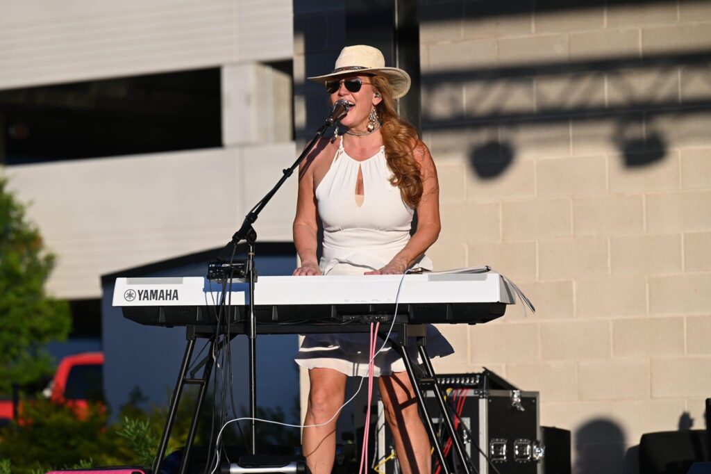 Dālene Richelle singing and playing keyboard outdoors in a white dress and cowboy hat.