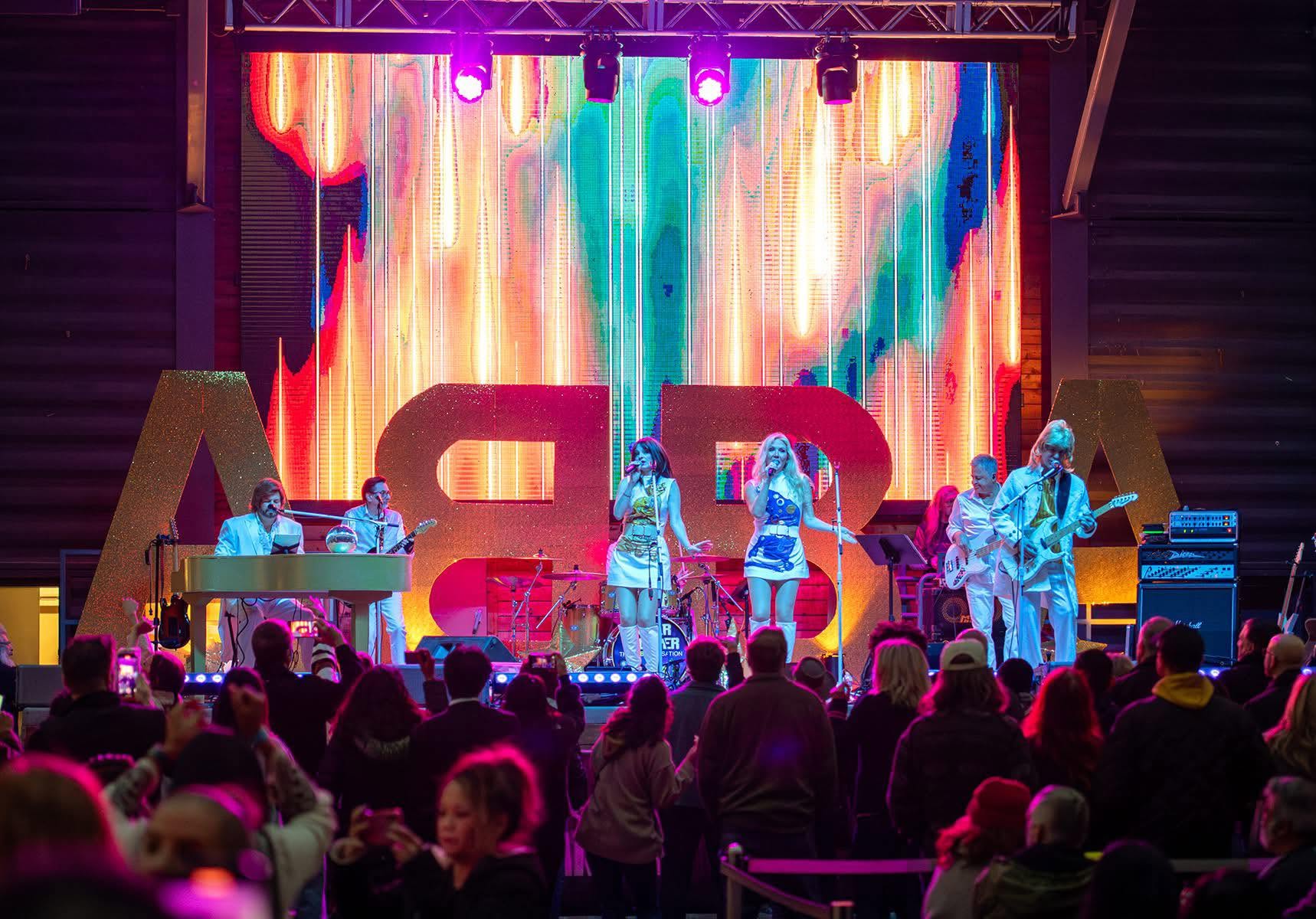 Super Trouper performing live with the full band before an audience and a large ABBA stage backdrop.