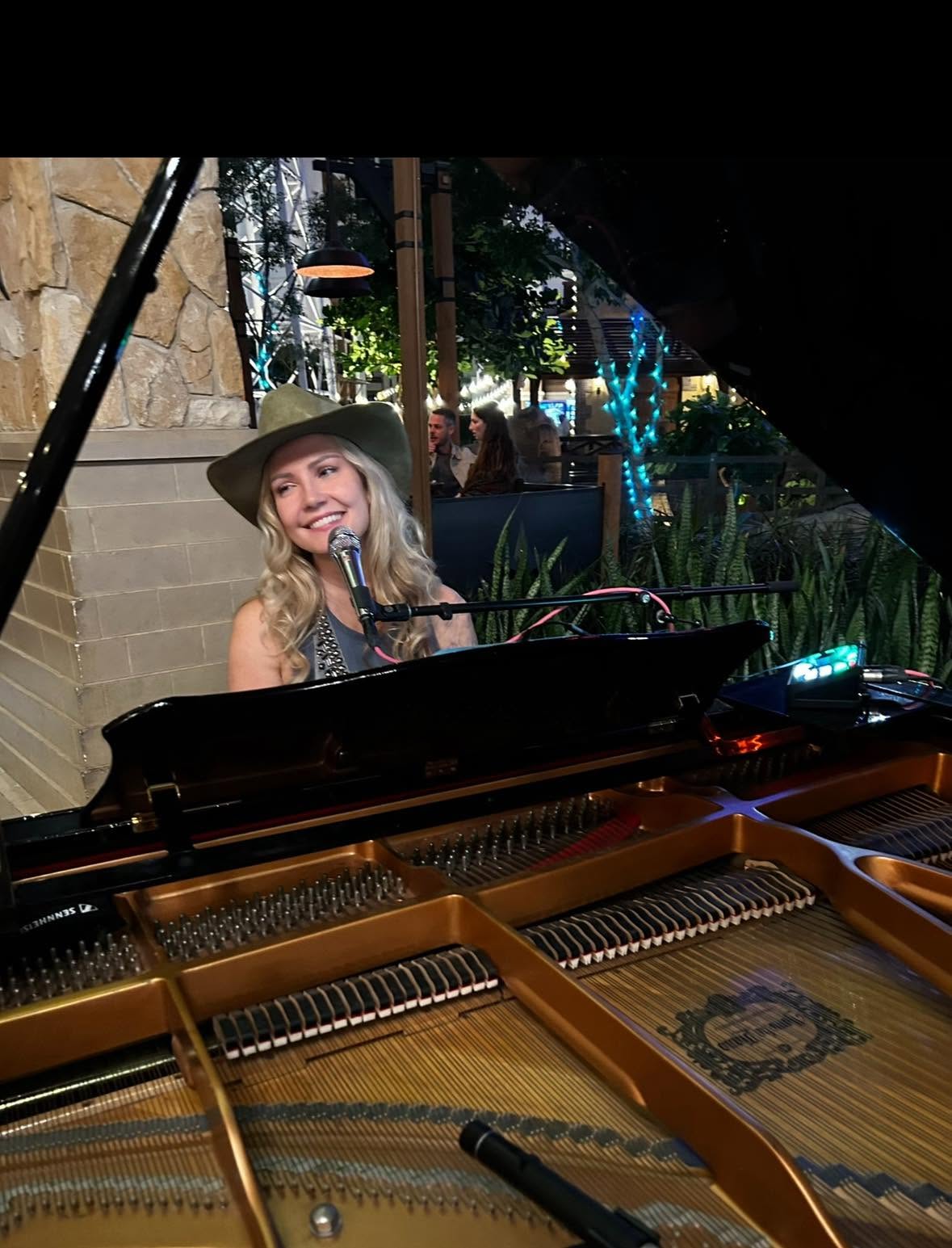 Dālene Richelle smiling through the open frame of a grand piano in a white cowboy hat.